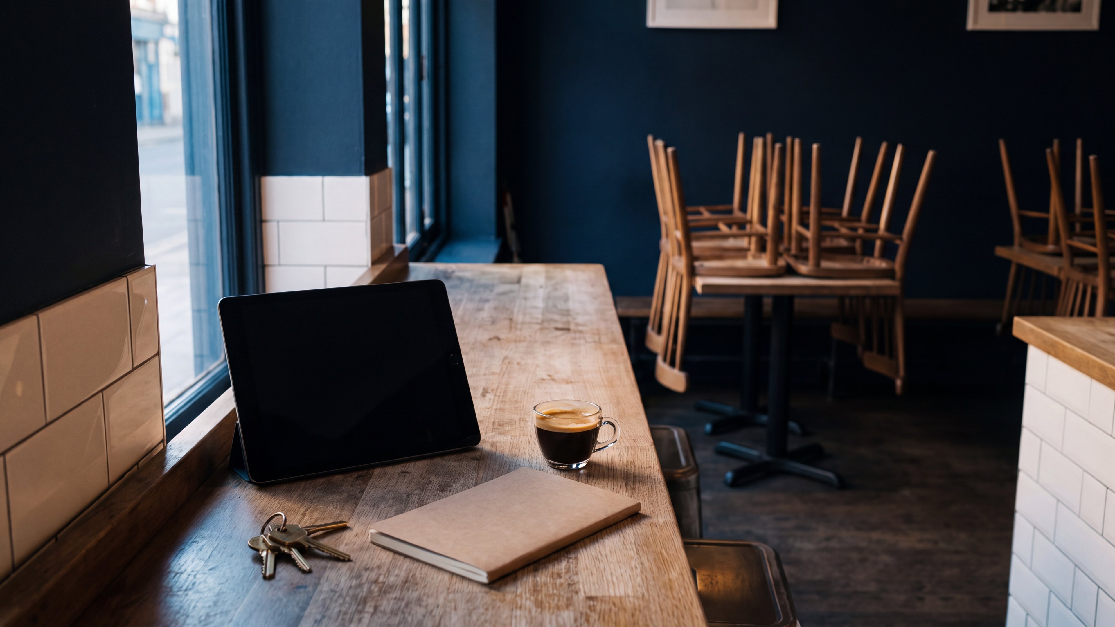 Kleines Café am frühen Morgen, Stühle noch auf den Tischen, auf der Theke ein Tablet, ein Espresso, ein Notizbuch und ein Schlüsselbund