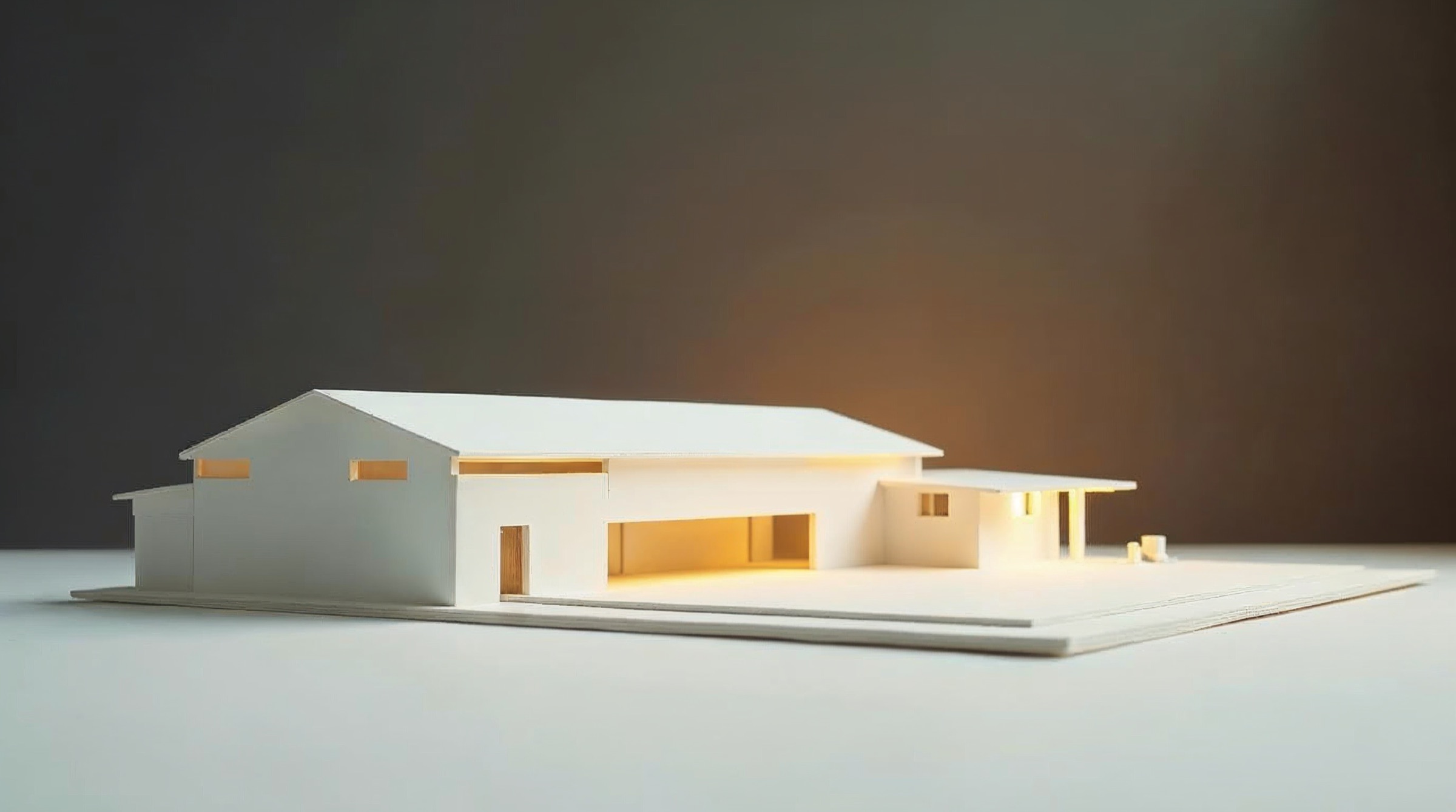 Ein weißes Architekturmodell eines Werksgebäudes, innen warm beleuchtet, auf heller Fläche vor dunklem Hintergrund.